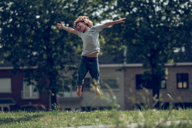 Todo el cuerpo de un alegre niño preadolescente descalzo saltando en una pradera verde en un día de verano en el campo