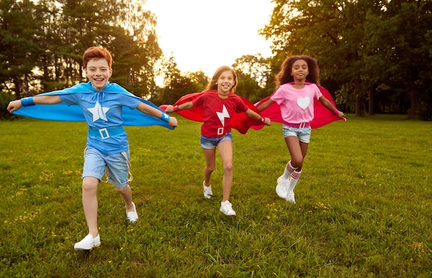Foto todo el cuerpo de alegres niños multirraciales en capas de superhéroe divirtiéndose y corriendo hacia la cámara en el prado verde en día de verano
