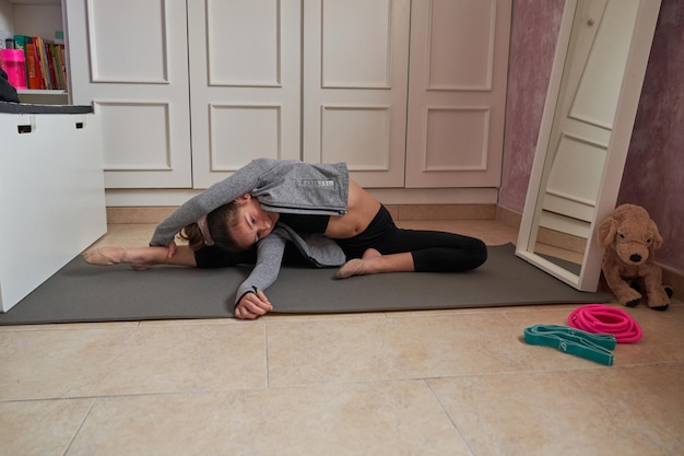 Todo el cuerpo de una chica concentrada, flexible y deportiva sentada sobre una alfombra gris y estirándose sola al lado del espejo