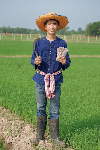 Todo el cuerpo del hombre agricultor asiático usa traje tradicional sonrisa permanente y sosteniendo dinero de billetes tailandeses en la granja verde