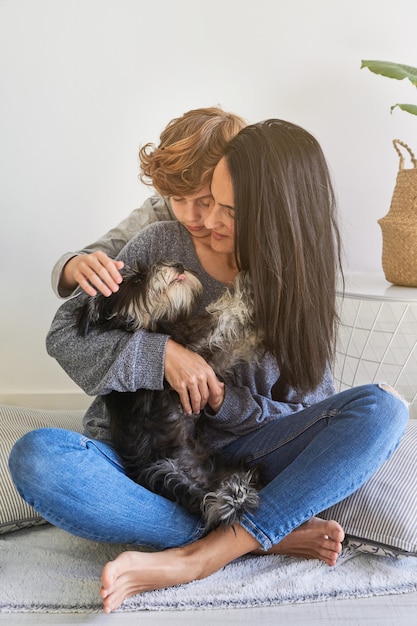 Foto todo el cuerpo de una joven de pelo largo con jeans y un niño de pelo rizado abrazando a un cachorro esponjoso y mirando a una mascota mientras estaba sentado en el suelo en una sala de estar moderna