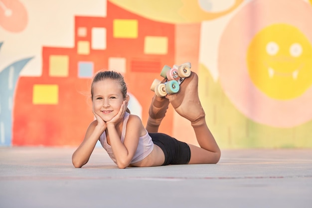 Foto todo el cuerpo de una joven sonriente en patines descansando sobre el asfalto y sosteniendo la cabeza entre las manos mientras mira la cámara