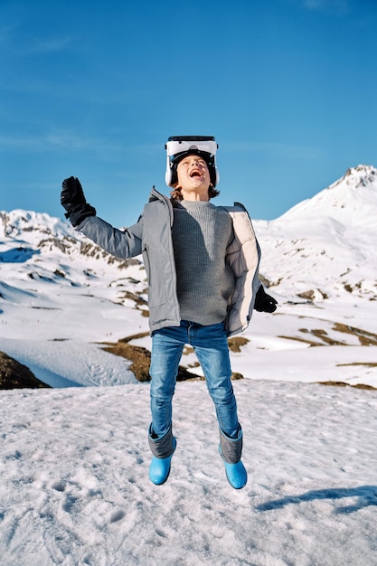 Todo el cuerpo de un niño feliz usando ropa exterior y auriculares VR saltando con el brazo levantado cerca de montañas nevadas bajo un cielo azul y mirando hacia arriba