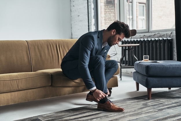 Todo debe ser perfecto. Apuesto joven en traje completo ajustando sus pantalones mientras está sentado en el sofá en casa