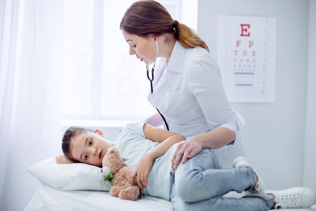 Todo está bien. Preocupado médico femenino hábil inclinado sobre la niña que miente y toca el oso de peluche