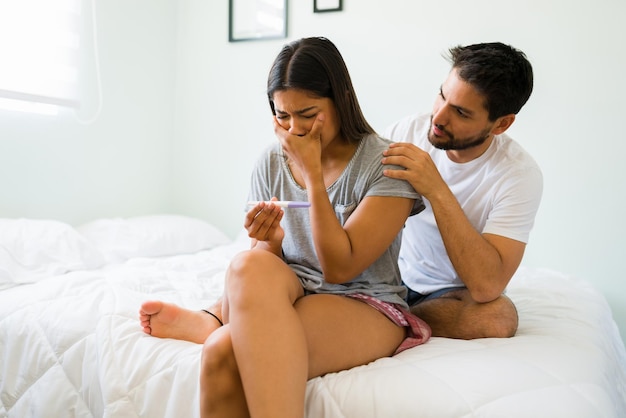 Todo va a estar bien. Mujer deprimida llorando lágrimas mientras mira su prueba de embarazo negativa mientras su pareja la abraza