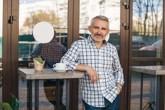 Todo excelente. Atractivo hombre de mediana edad sonriente mirando con confianza a la cámara de pie de espaldas al café al aire libre