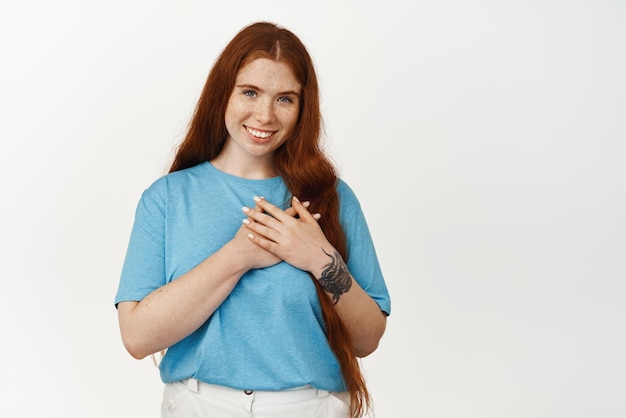 Con todo mi corazón, una joven pelirroja sonriente se toma de la mano en el pecho, se ve halagada y agradecida, dice gracias, aprecia algo de pie contra el fondo blanco