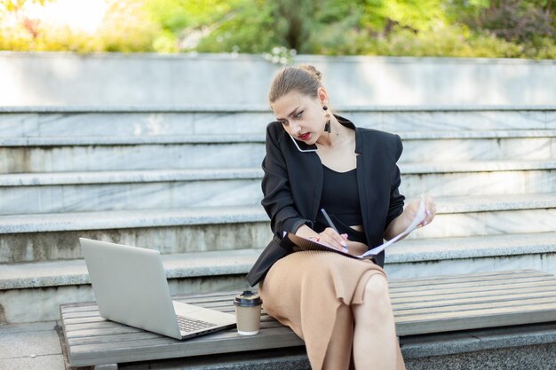 Todo en una mujer de negocios hablando por teléfono escribiendo en una carpeta con papel sentado en un banco con una computadora portátil al aire libre