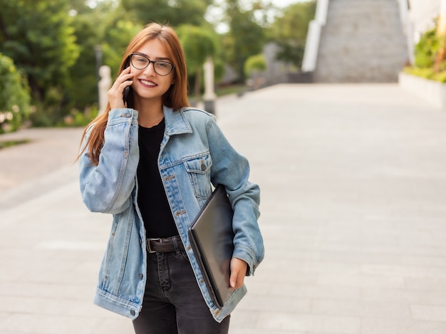 Todo en el negocio. Mujer elegante joven en una chaqueta de mezclilla y gafas sosteniendo portátil y hablando por teléfono mientras camina en la ciudad.