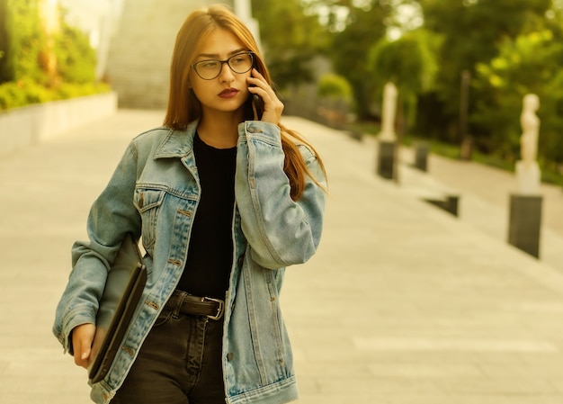 Todo en el negocio. Mujer joven con estilo en una chaqueta de mezclilla y gafas sosteniendo portátil y hablando por teléfono mientras camina por la ciudad.