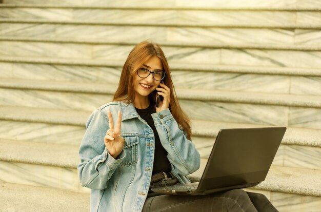 Todo en el negocio. Mujer joven con estilo en una chaqueta de mezclilla y gafas usa una computadora portátil y hablando por teléfono mientras está sentado en las escaleras de la ciudad. Trabajo remoto.