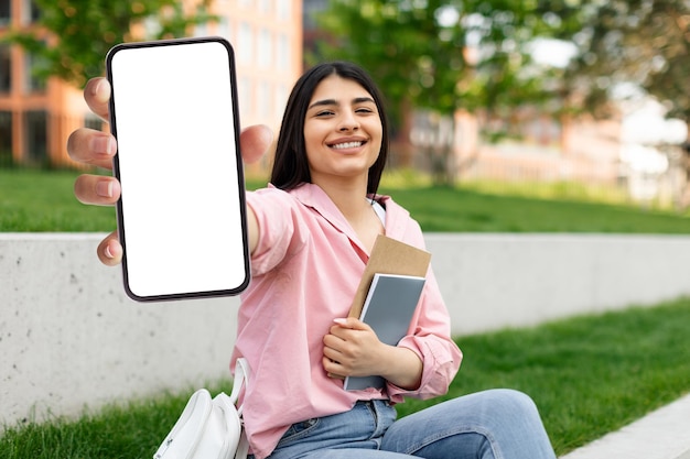 Tolle Lern-App. Glückliche Studentin zeigt im Sitzen ihr Smartphone mit leerem Bildschirm
