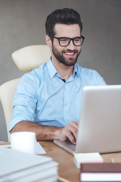 Tolle Lösung. Nahaufnahme fröhlicher junger gutaussehender Mann mit Brille, der am Laptop arbeitet und lächelt