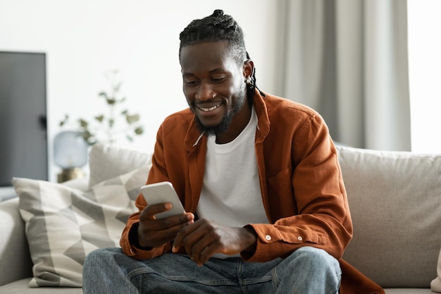 Tolle mobile App Positiver schwarzer Mann, der Handy-SMS verwendet und im Internet surft, sitzt auf dem Sofa zu Hause freier Speicherplatz