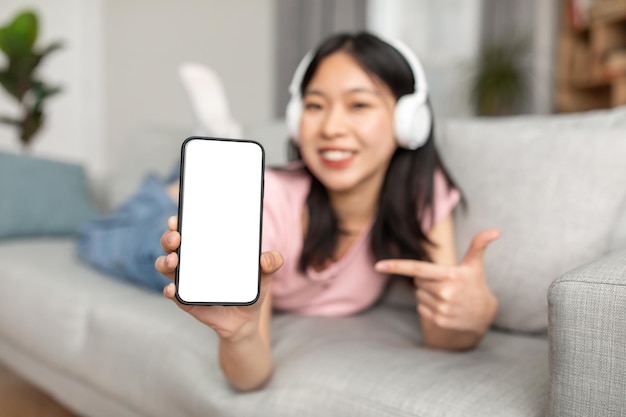 Tolle musikalische App, glückliche asiatische Dame, die einen leeren Bildschirm des Telefons mit drahtlosen Kopfhörern zeigt, die aufliegen