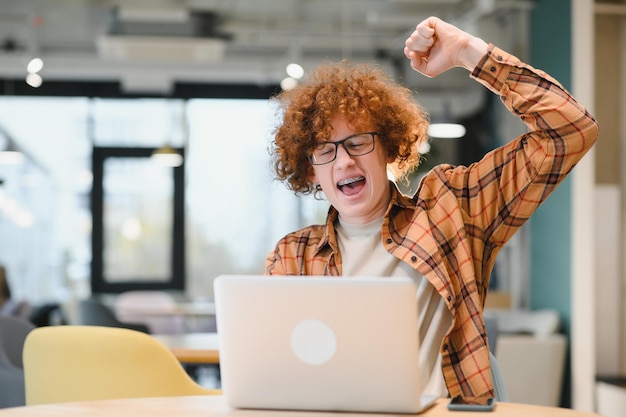 Foto tolle neuigkeiten männlicher teenager drückt erfolg vor dem laptop im café aus, ballt seine faust und schreit kopienraum