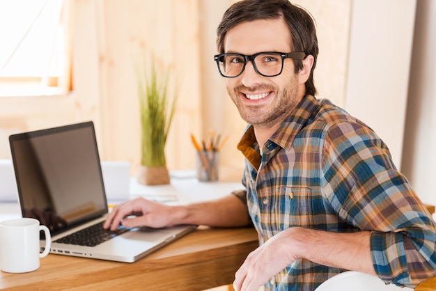 Tolle Stimmung um gut zu arbeiten. Schöner junger Mann, der an seinem Laptop arbeitet und die Kamera über die Schultern schaut, während er an seinem Arbeitsplatz sitzt