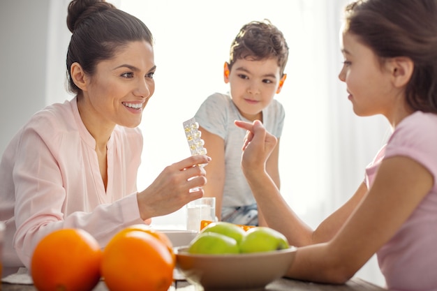 Tolle Vitamine. Nette überschwängliche dunkelhaarige junge Mutter, die Vitamine hält und mit ihren Kindern über das Gesundheitswesen und den Jungen spricht, der auf dem Tisch sitzt