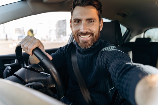 Foto tolles selfie. hübscher bärtiger mann, der kamera hält und selfie macht, während er im auto auf dem fahrersitz sitzt und die kamera mit freude anlächeln