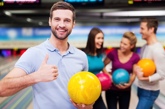 Tolles Spiel! Hübsche junge Männer, die eine Bowlingkugel und den Daumen hochhalten, während drei Leute gegen Bowlingbahnen kommunizieren