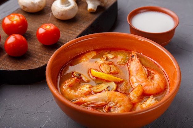 Tom Yam Suppe mit Garnelen in einer Terrine auf einem konkreten Hintergrund neben einer Schüssel mit Kokosmilchpilzen und Tomaten. Horizontales Foto