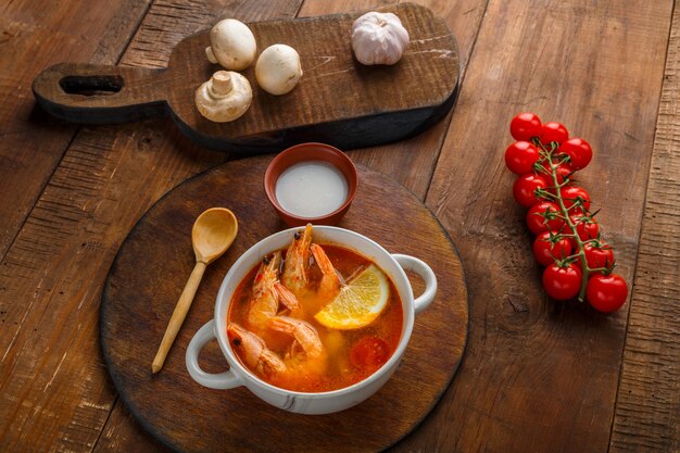 Tom Yamsuppe mit Garnelen und Kokosmilch auf dem Tisch auf einem runden Brett neben Kokosmilchpilzen und Tomaten.