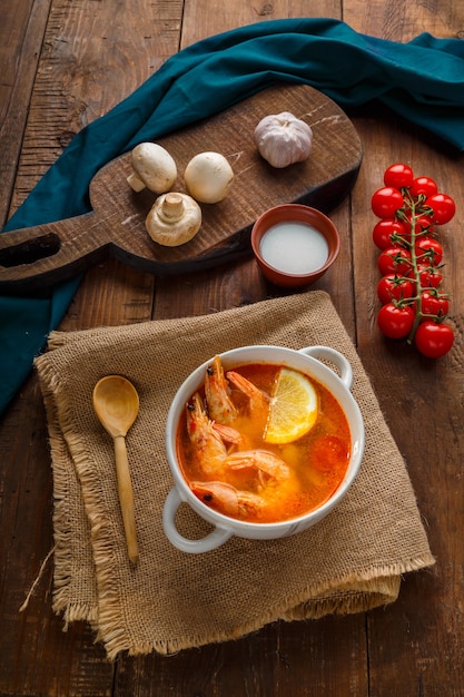 Tom Yamsuppe mit Garnelen und Kokosmilch auf einem Tisch auf Holzbrettern in der Nähe von Milchpilzen und Tomaten. Vertikales Foto