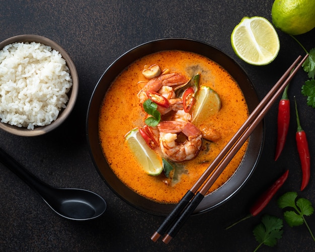 Tom Yum Suppe mit Garnelen in einer schwarzen Keramikschale mit Stäbchen auf dunklem Hintergrund Draufsicht dark