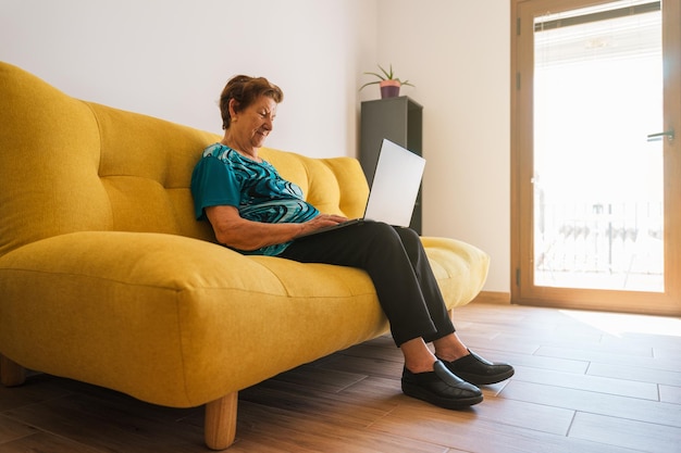 Toma abierta de una anciana caucásica que usa una computadora portátil en un sofá amarillo. Lleva una camiseta azul.