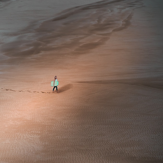Toma aérea joven sosteniendo una tabla de surf caminando en un desierto vacío dejando huellas de sus pies