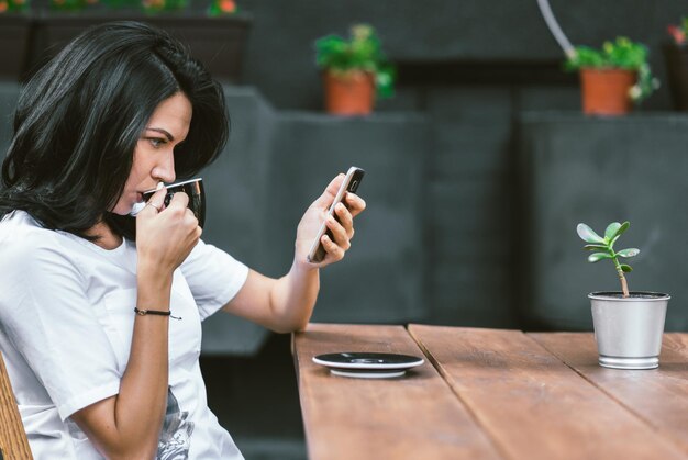 Toma al aire libre de una chica caucásica con camiseta blanca disfrutando de wifi gratis en una cafetería navegando por Internet en un teléfono móvil leyendo noticias y bebiendo café por la mañana Gente y concepto de estilo de vida