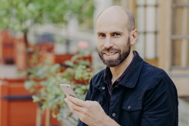 Toma al aire libre de un hombre apuesto con barba oscura y bigote que está de buen humor, pasa el tiempo libre afuera y sostiene búsquedas de teléfonos inteligentes en las redes sociales conectadas a Internet inalámbrico