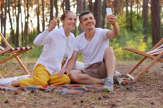 Toma al aire libre de una pareja sonriente sentada en el bosque y transmitiendo en vivo o haciendo videollamadas marido y mujer haciendo selfie mujer mostrando v signo hombre tomando fotos con teléfono inteligente