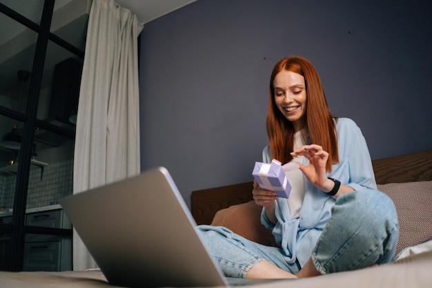 Toma de ángulo bajo de una mujer sonriente abriendo una caja de regalo con un regalo mientras realiza una videollamada en una computadora portátil