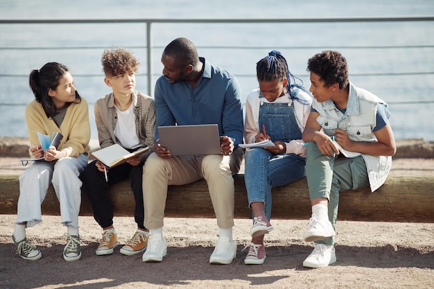 Toma completa de un grupo diverso de niños con un maestro que usa una computadora portátil al aire libre durante la escuela de verano