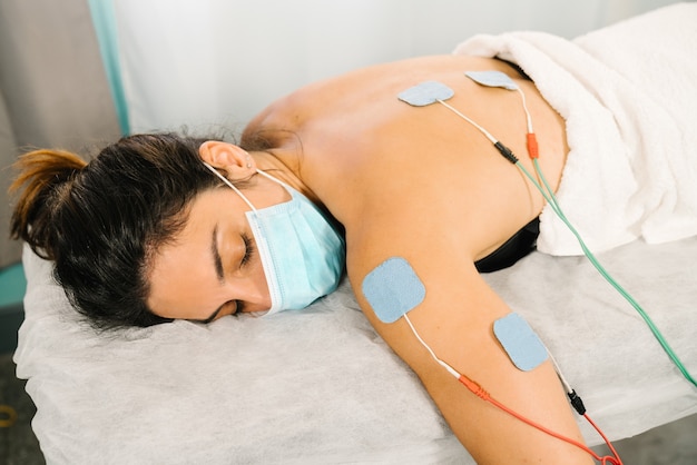 Foto toma corta de una mujer caucásica que recibe tratamiento de fisioterapia con electrodos de electroestimulación en la espalda mientras está acostada en una camilla con una mascarilla debido a la pandemia del coronavirus covid 19.