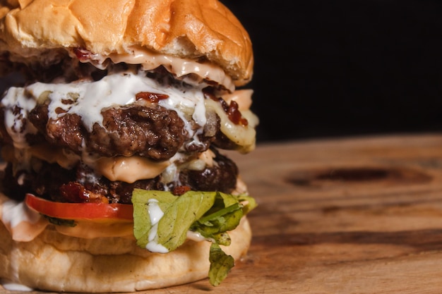 Toma detalle de una hamburguesa doble gigante con condimentos encima de una mesa de madera con fondo negro.