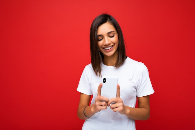 Toma de la foto de atractiva mujer joven de buen aspecto positivo vistiendo ropa casual elegante poising aislado sobre fondo con espacio vacío sosteniendo en la mano y utilizando sms de mensajería de teléfono móvil mirando