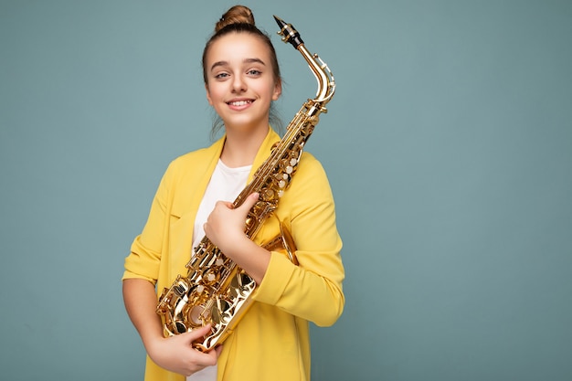 Toma de la foto de la atractiva niña morena sonriente positiva vistiendo chaqueta amarilla de moda que se encuentran aisladas sobre fondo azul pared sosteniendo el saxofón mirando a la cámara