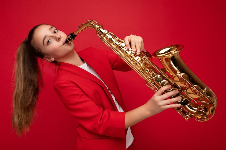 Foto toma de la foto de la hermosa dama morena positiva feliz vistiendo elegante chaqueta roja que se encuentran aisladas sobre la pared de fondo rojo tocando el saxofón mirando a la cámara