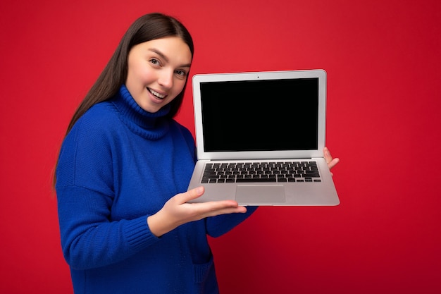 Toma de la foto de la hermosa y encantadora mujer morena joven sonriente sosteniendo el ordenador portátil mirando a la cámara en suéter azul aislado sobre fondo de pared roja. Espacio vacio