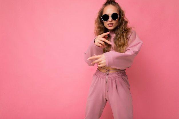 Toma de la foto de la hermosa joven rubia oscura feliz vistiendo ropa casual y elegantes gafas de sol aisladas sobre fondo de colores mirando a la cámara y divirtiéndose