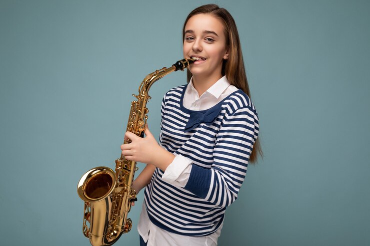 Foto toma de la foto de la hermosa niña morena sonriente feliz con rayas de manga larga que se encuentran aisladas sobre la pared de fondo azul tocando el saxofón mirando a la cámara.