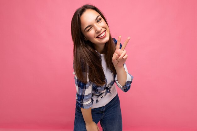 Toma de la foto de la joven mujer bonita morena sonriente encantadora positiva con emociones sinceras con camisa a cuadros hipster que se encuentran aisladas sobre fondo rosa con espacio libre y mostrando el signo de la paz.