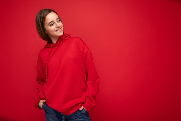 Toma de la foto de la niña morena sonriente bastante positiva vistiendo elegante sudadera con capucha roja de pie