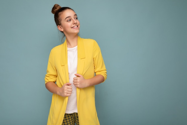 Toma de la foto de la niña morena sonriente feliz bastante positiva que lleva la chaqueta amarilla de moda y la camiseta blanca que se encuentran aisladas sobre la pared de fondo azul mirando hacia el lado. Copia espacio