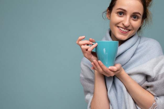 Toma de la foto del primer de la mujer morena sonriente feliz joven hermosa que lleva la bufanda azul caliente aislada sobre fondo azul que sostiene la taza azul que bebe café y que mira la cámara. copia espacio