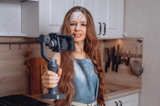 Toma de fotografías para redes sociales. Una joven atractiva sostiene un teléfono con un trípode y posa para un selfie. Foto en el estilo culinario. Un delantal de casa azul y harina por toda la cara.