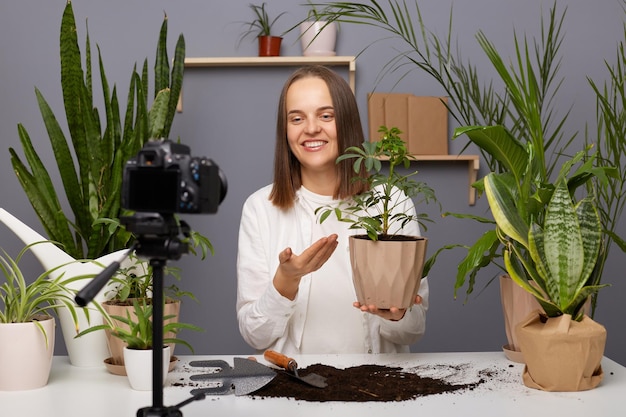 Toma horizontal de una bloguera con cabello castaño blogueando sobre plantas de interior usando una cámara en un trípode estabilizador para grabar un video en su vlog sosteniendo y mostrando una maceta con flores